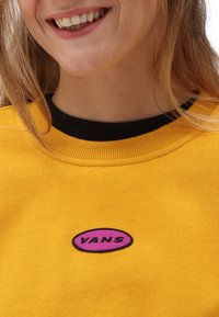 Žlutá mikina s texturovaným černým límcem a vyšitou oválnou nášivkou v růžové a černé barvě, na které je zobrazeno jméno značky "VANS."
