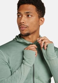 Uomo con capelli ricci corti che indossa un hoodie con zip di colore verde chiaro, mentre tira su la zip guardando di lato.