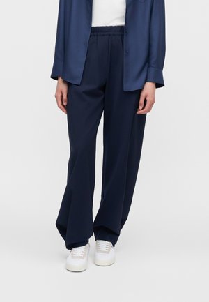 Persoon die marineblauwe losse broek, witte sneakers, een wit topje en een marineblauw overhemd met knopen draagt, staand tegen een effen achtergrond.
