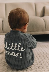 Cardigan lavorato a maglia di un grigio scuro con una texture a coste, con la scritta ricamata in bianco "little man" sulla schiena, seduto su un tappeto texture.