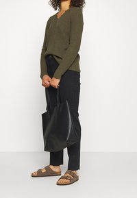 Olivgrüner gerippter Pullover mit gewelltem Ausschnitt, schwarze hochgeschnittene Jeans, braune Sandalen und eine große schwarze Tragetasche.