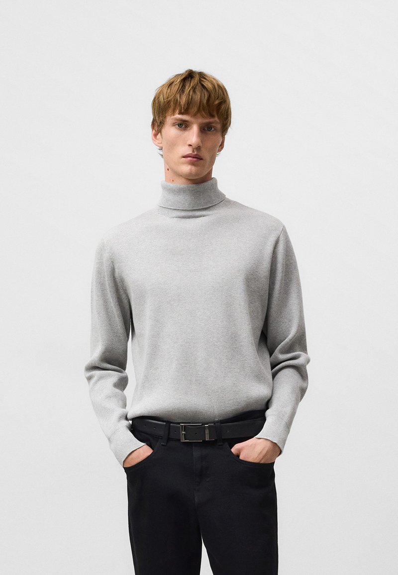 Szary sweter z golfem o prążkowanej fakturze, z długimi rękawami i kołnierzem średnio-wysokim, zestawiony z czarnymi spodniami i paskiem.