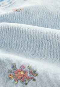 Tissu en denim bleu clair avec un tissage texturé, mettant en avant une broderie florale en rose, jaune et vert sur le bord pour un détail décoratif.