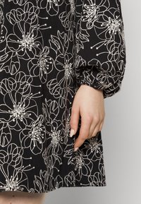 Robe noire ornée de broderies florales blanches, avec des manches bouffantes et une coupe décontractée. Texture lisse avec des détails de couture visibles.