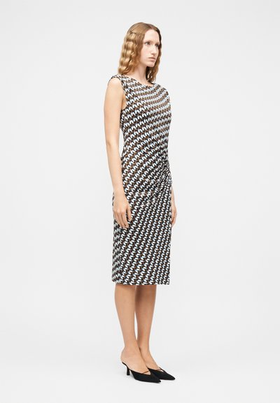 Γυναίκα με κυματιστά μαλλιά, που φορά αμάνικο φόρεμα μήκους μέχρι το γόνατο με γεωμετρικό μοτίβο σε μπλε και καφέ αποχρώσεις και μαύρες μυτερές γόβες, όρθια πλάγια.