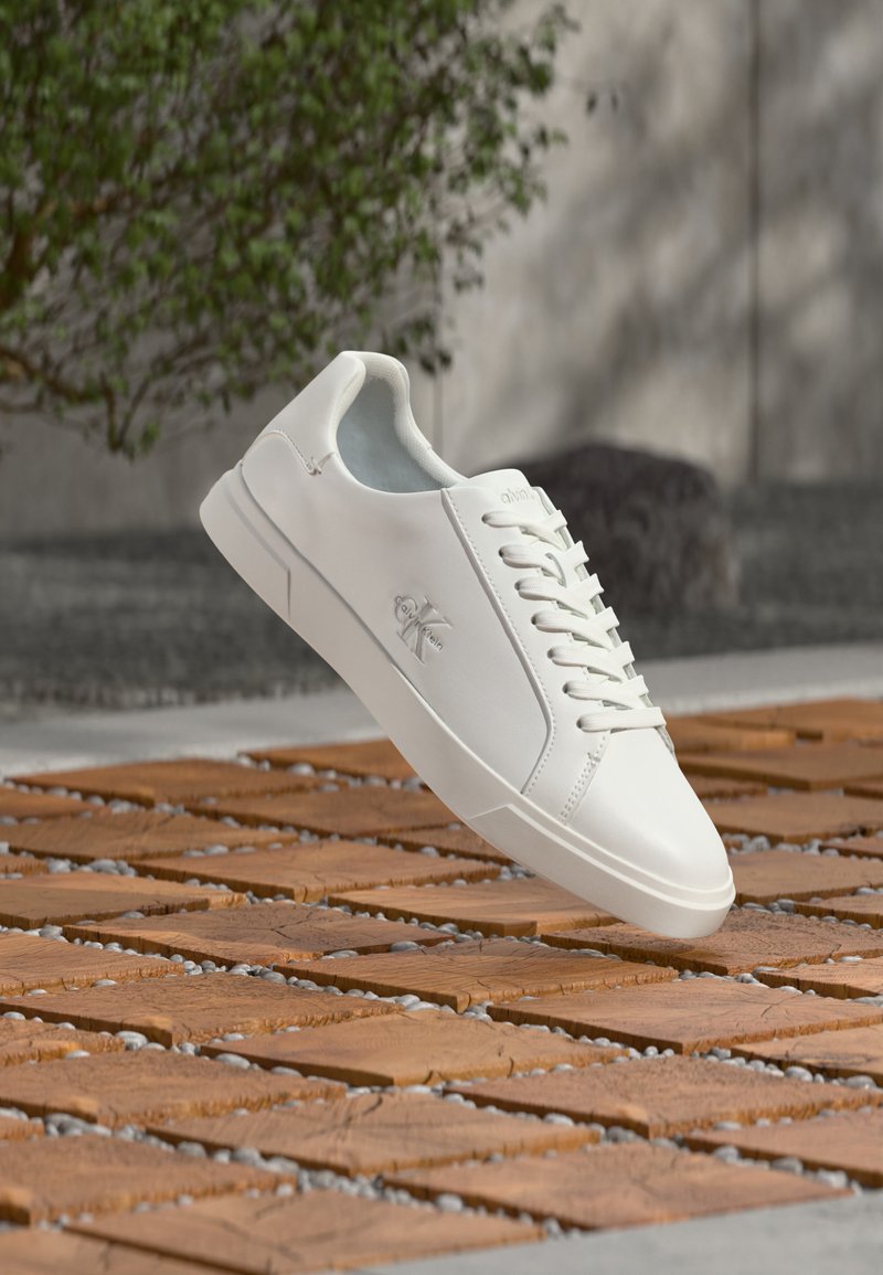 Λευκό χαμηλό αθλητικό παπούτσι με κορδόνια και λογότυπο Calvin Klein στο πλάι, αιωρείται πάνω από ξύλινη πλακόστρωτη επιφάνεια έξω, κοντά σε πράσινο.