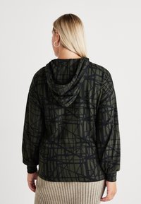 Hooded sweatshirt v temno zeleni barvi z črnimi angularnimi vzorci, dolge rokave in sproščen kroj. Teksturni material z rebrastimi detajli.