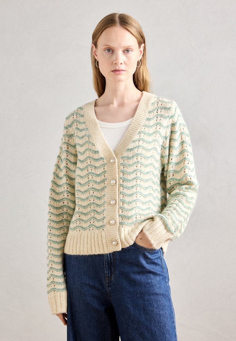 Cardigan de malha creme com um padrão de ondas em verde claro. Apresenta punhos e barra canelados, e cinco botões de pérola na parte da frente.