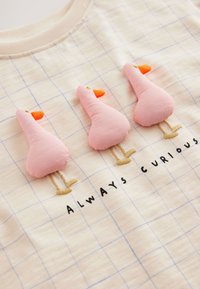 Bomuld T-shirt med lys beige baggrund med blåt ternmønster, tre lyserøde stoffugle og broderet tekst "ALWAYS CURIOUS."