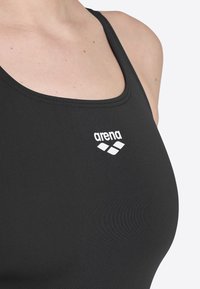 Schwarzer Badeanzug aus dehnbarem Material mit einem runden Ausschnitt und einem weißen Logo auf der Brust. Glatte Textur und taillierter Schnitt.
