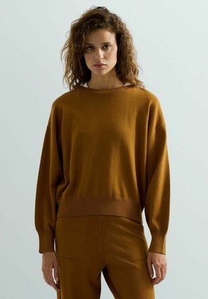 Donna con capelli ricci che indossa un maglione marrone largo e pantaloni coordinati, in piedi davanti a uno sfondo chiaro e semplice.