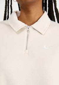 Φωτεινό μπεζ πλεκτό πόλο ζακέτα με φερμουάρ και γιακά, που διαθέτει ένα μικρό λευκό λογότυπο Nike στο στήθος και απαλή υφή από μείγμα βαμβακιού.