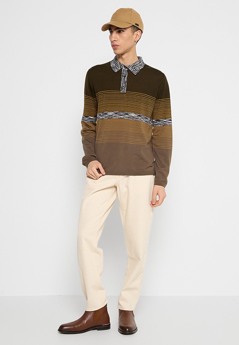 Langarm-Polohemd in braun-gelben Streifen mit einem gemusterten schwarzen Kragen. Kombiniert mit cremefarbigen Hosen und braunen Stiefeletten.