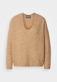 Pull en maille beige à manches longues avec un col en V profond et un petit logo métallique rond près de l'ourlet inférieur.