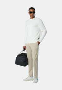Maglione bianco a coste, pantaloni beige, borsa duffle nera e sneakers bianche. Modello con occhiali da sole, in piedi contro uno sfondo neutro.