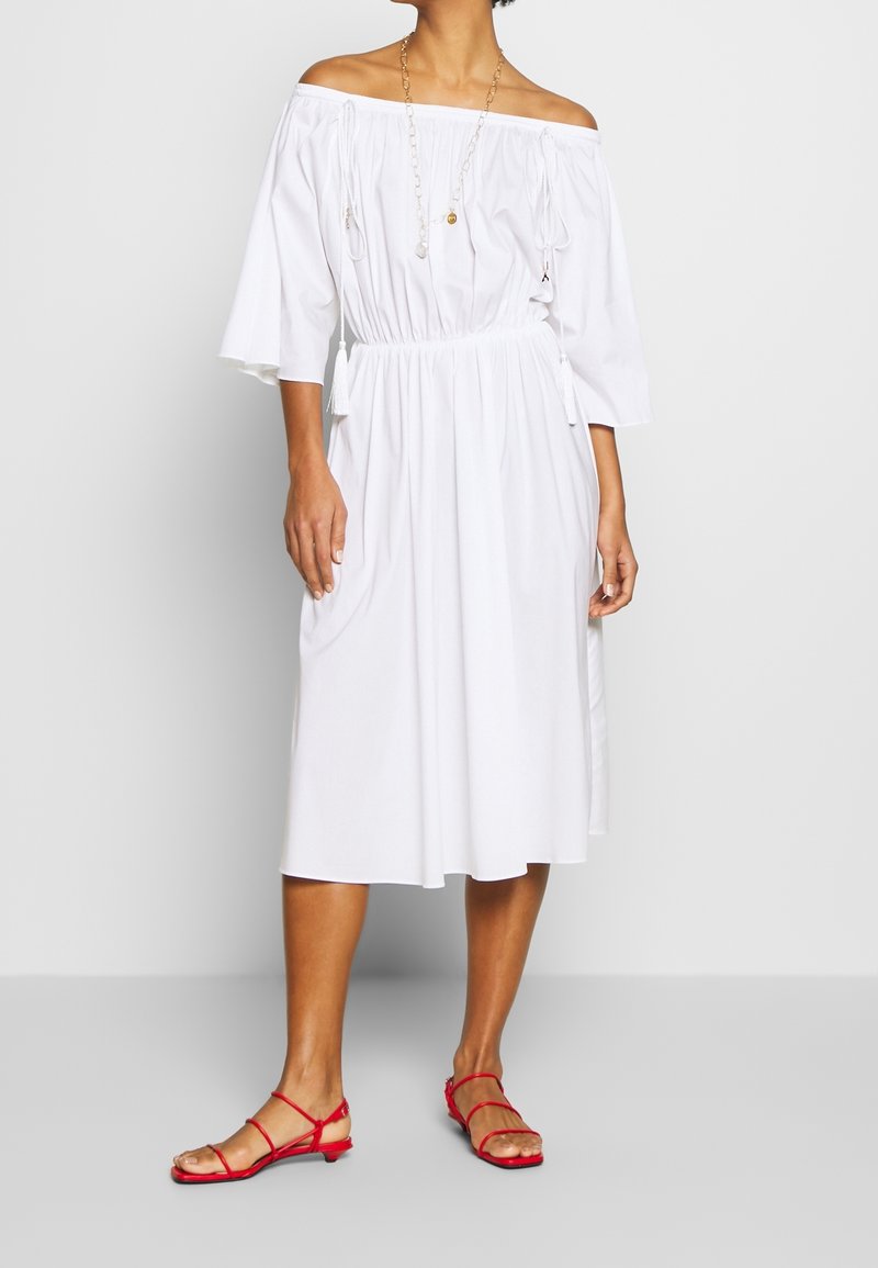 Femme portant une robe midi blanche à épaules dénudées avec des manches 3/4, une taille cintrée, un long collier et des sandales plates rouges à lanières, debout sur un sol gris.