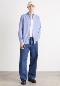 Niebiesko-biała koszula w paski zapinana na guziki, pod spodem biały top na ramiączkach, luźne niebieskie dżinsy, czarne buty i beżowa czapka. Prosty design, swobodny krój.