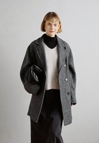 Abrigo de lana gris con diseño de doble botonadura, combinado con un suéter de punto ligero y jeans negros de tiro alto. Acento con bolso de mano negro.