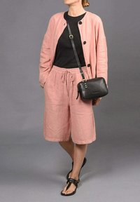 Cárdigan de lino rosa claro con botones negros sobre una camiseta negra, combinado con pantalones cortos rosa a juego y sandalias negras. Bolso bandolera negro.