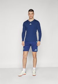 Camisa atlética de manga larga azul con cuello redondo, combinada con pantalones cortos azules que presentan un logo y acentos en azul claro. Zapatillas deportivas blancas.