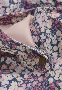 Blommönstrad tyg med rosa, vita och marinblå blommor, med en mjuk foder och en detaljerad mässingsknapp vid kragen.