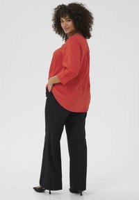 Blouse rouge à manches longues avec une finition texturée, associée à un pantalon noir ample et des talons aiguilles noirs. Détail d'ourlet arrondi visible.