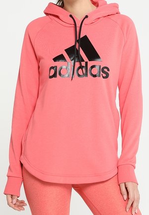 Kvinde iført en koralpink Adidas-hoodie med sort logo og matchende koralpink leggings, stående med hænderne afslappet ved siden.