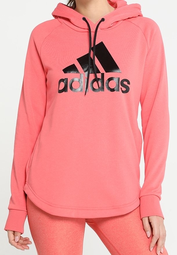 Femme portant un hoodie corail rose Adidas avec un logo noir et des leggings corail rose assortis, debout les mains détendues le long du corps.