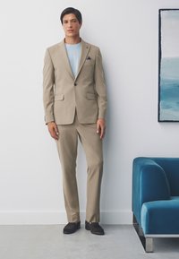Costume beige à coupe ajustée, avec une fermeture à un bouton et des revers crantés. Associé à une chemise bleu clair et des mocassins foncés.