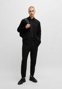 Zwarte verstelbare jas en bijpassende joggers, beide met een gladde textuur. De jas heeft een gele logo; de joggers hebben elastische boorden en een trekkoord.