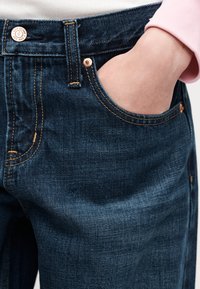 Χέρι στην μπροστινή τσέπη σκούρου μπλε τζιν με χαλκοκολήματα και χρυσό κουμπί, φορεμένα με ροζ ανοιχτόχρωμο μπλουζάκι με μακριά μανίκια.