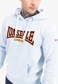 Felpa con cappuccio azzurro chiaro, dotata di una tasca frontale e cordini bianchi. Presenta il logo "Lonsdale London" in bordeaux e giallo sul petto.