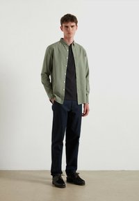 Lichtgroen overhemd met knopen over een zwart T-shirt, gecombineerd met marineblauwe broek en donkere schoenen. Het heeft een kraag en knopen aan de voorkant.