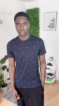 Mornarica majica kratkih rukava s bijelim uzorcima strelica i džepom na prsima, u kombinaciji s crnim hlačama. Prostorija sadrži zelenilo i bijelu policu.