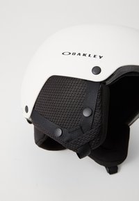 Biały kask narciarski z czarnym wyściełaniem, charakteryzujący się teksturą siatki i regulowanymi paskami. Logo nadrukowane na górze, akcentowane czarnymi guzikami.