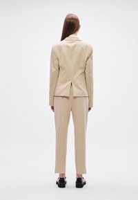 Béžový oblek na mieru so priliehavým sakom a rovným nohavicami. Textúrovaná látka, detail rozparku na zadnej strane, skombinované s čiernymi topánkami.