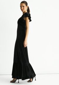 Black velvet maxi dress with ruffled short sleeves, fitted bodice, and flared hem. Paired with black heeled sandals.