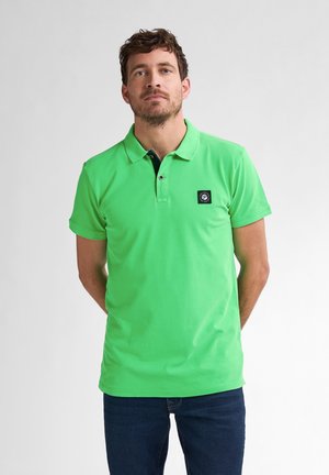 Polo de color verde brillante hecho de algodón suave con un cuello negro, que presenta un parche de logo en el lado izquierdo del pecho y mangas cortas.