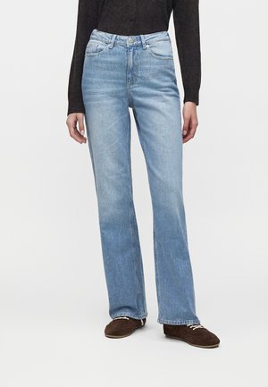 Personne portant un jean droit bleu clair, un haut à manches longues foncé et des chaussures marron foncé avec des lacets blancs, debout devant un fond uni.