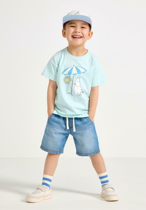 Smilende dreng iført en lyseblå kasket, t-shirt med en tegneserie og paraply, denimshorts, stribede sokker og hvide sneakers, stående med hænderne i lommerne.