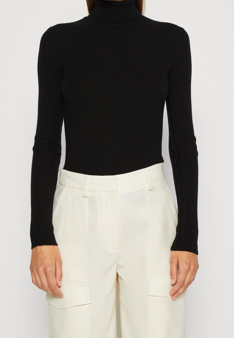 Personne portant un pull à col roulé côtelé noir et un pantalon blanc taille haute à jambes larges avec poches cargo, debout devant un fond uni.