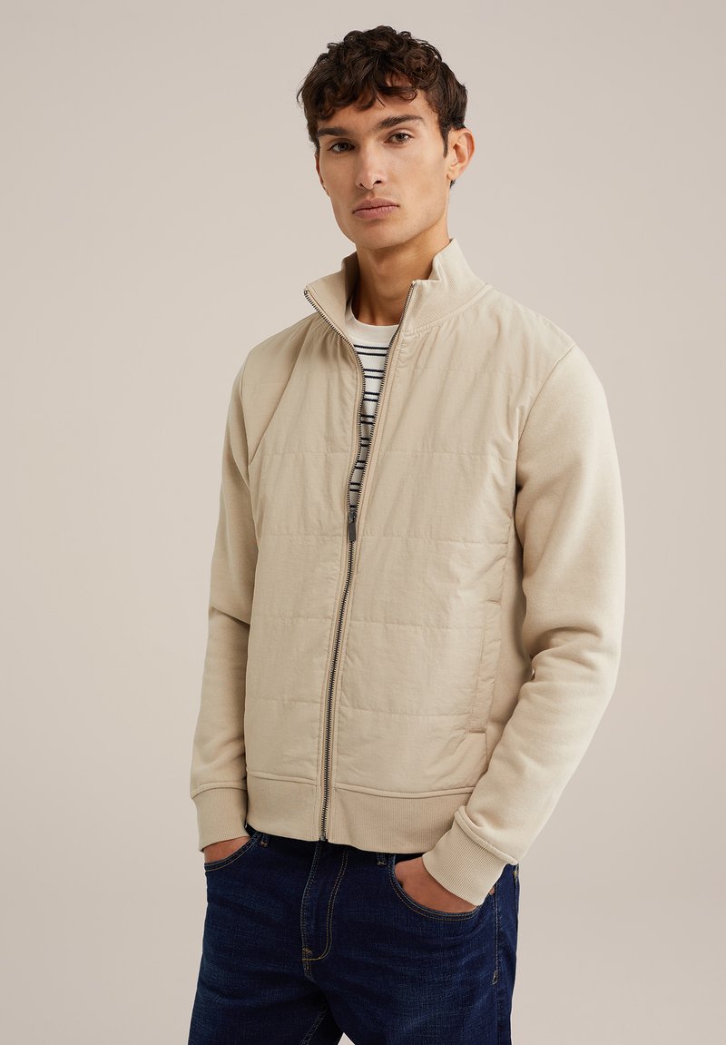 Giacca beige con zip, frontale imbottito, maniche a maglia e colletto alto. Indossata sopra una camicia a righe, abbinata a jeans blu scuri.