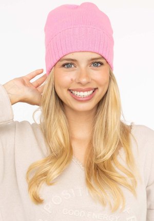 Pink knitted beanie with a ribbed texture and a folded cuff, worn by a smiling person with long, straight blonde hair.