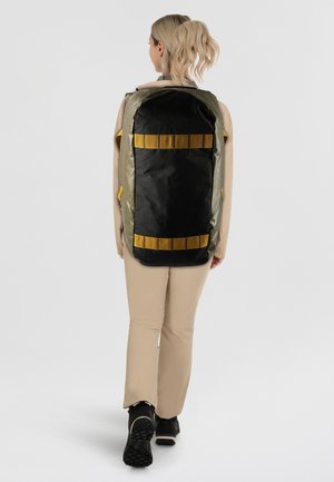 Mochila con un cuerpo de tela negra y acentos en verde oliva, que presenta correas y cremalleras amarillas. Material texturizado, forma rectangular.