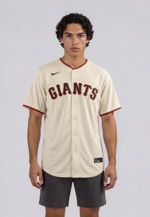 Jonge man met krullend haar die een beige Giants honkbalshirt en donkere korte broek draagt, staand tegen een effen lichtgrijze achtergrond.