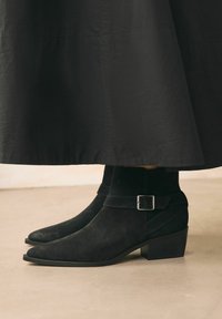 Botines de ante negros con punta afilada, un ligero tacón y un detalle de correa con hebilla decorativa. Sobre una falda negra en una superficie neutra.
