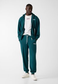 Turkooizen trainingspak gemaakt van zacht materiaal, met een jas met ritssluiting en zijzakken en een logo, gecombineerd met bijpassende broek en witte sportschoenen.