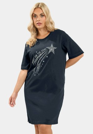 Lyshåret kvinde iført en løs, sort kjole med korte ærmer og en glitrende stjerneskudsgrafik med "Shoot for the stars" tekst.