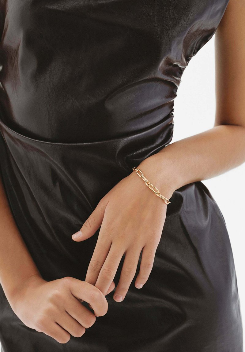 Pulsera de cadena de oro en una mano, sobre un vestido de cuero negro. Metal suave con eslabones interconectados, mostrando un acabado pulido.