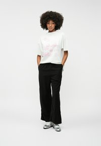 Fehér, túlságosan nagy póló rózsaszín grafikai mintával, fekete, laza fazonú nadrággal és ezüst lapos cipővel párosítva. Simán textúrák, kényelmes szabás.